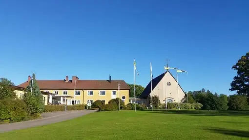 Huvudbyggnaden och kyrkan vid Flämslätt Stifts- och Kursgård, med gröna gräsmattor och svenska flaggor.