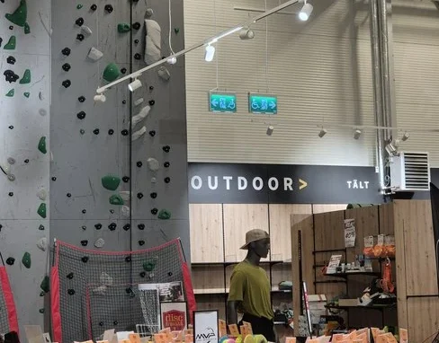 Astro Sweden - Klätterväggen Hög klättervägg placerad i ett hörn av Astro Sweden, omgiven av outdoorprodukter.
