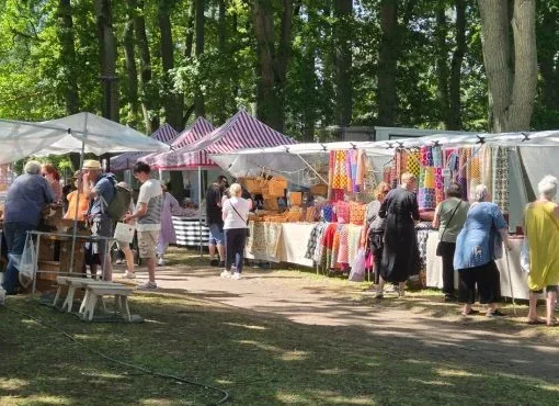 Hantverksmässa utomhus med människor som tittar på handgjorda produkter vid marknadsstånd, omgivna av träd och färgglada tygdekorationer.