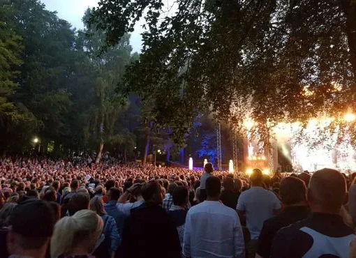 Stor publik samlad framför en utomhusscen med livemusik, ljusshow och träd runtom, en stämningsfull musikfest med dans och gemenskap.